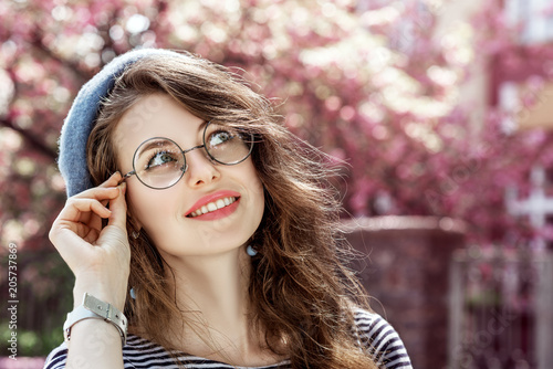 Outdoor close up portrait of young beautiful happy smiling girl wearing round transparent glasses, blue beret. Model looking up, posing in street. Spring fashion concept. Copy, empty space for text
