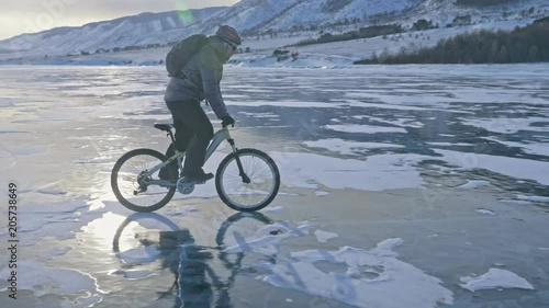 Man is riding a bicycle on ice. The cyclist is dressed in a gray down jacket, backpack and helmet. Ice of the frozen Lake Baikal. The tires on the bicycle are covered with special spikes. The traveler photo