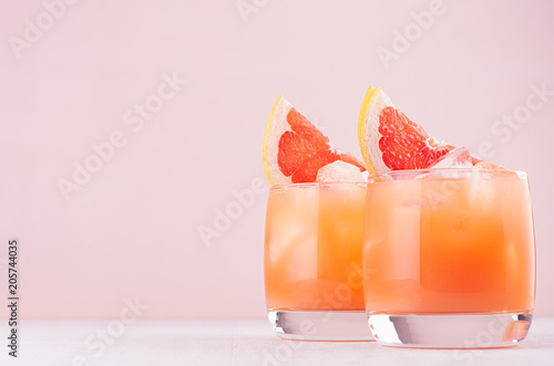 Cold grapefruit cocktail in two glasses with ice and pieces grapefruit on pastel pink background. Fresh summer healthy diet beverage.