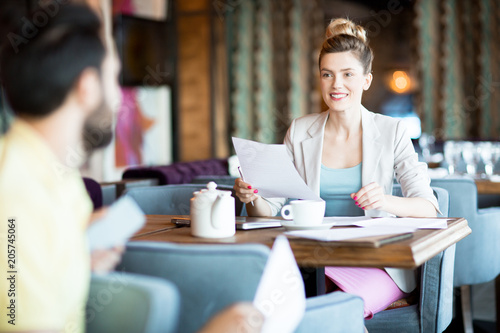 Happy young businesswoman with paper sitting by table in cafe, looking at colleague and discussing it with him