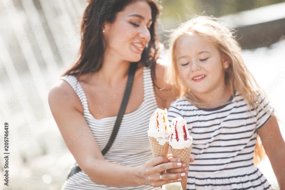 Young pretty mother and her daughter having fun together near the fountain. Beautiful woman and her little child eating ice cream. Cheerful family having fun. Young pretty mother and her daughter having fun together near the fountain. Beautiful woman and her little child eating ice cream. Cheerful family having fun.