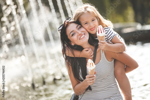 Young pretty mother and her daughter having fun together near the fountain. Beautiful woman and her little child eating ice cream. Cheerful family having fun.