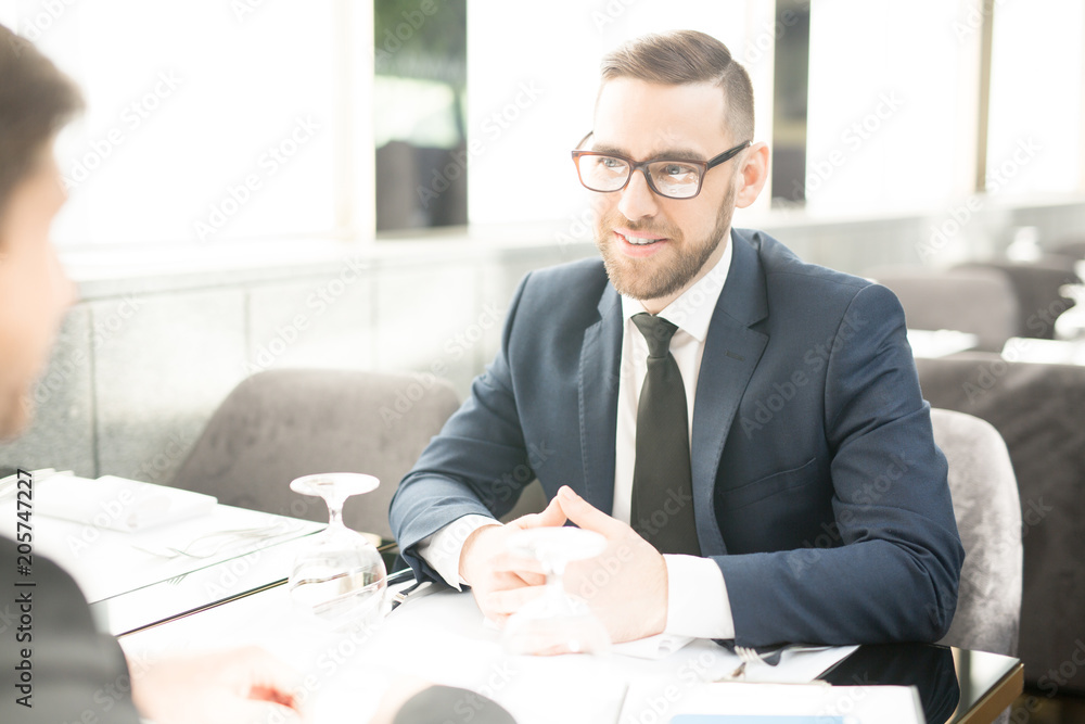 Confident businessman in suit and eyeglasses negotiating with one of entrepreneurs in cafe or restaurant Confident businessman in suit and eyeglasses negotiating with one of entrepreneurs in cafe or restaurant