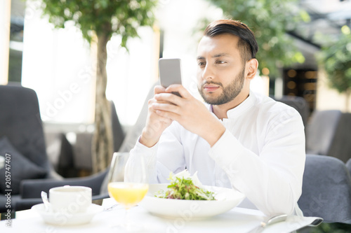 Young businessman sitting in cafe with fresh vegetarian salad and drinks on table and texting in smartphone