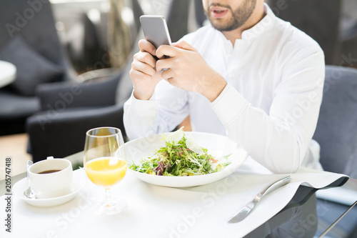 Fototapeta Naklejka Na Ścianę i Meble -  Young entrepreneur texting in his smartphone over served table with fresh salad, glass of orange juice and cup of black coffee