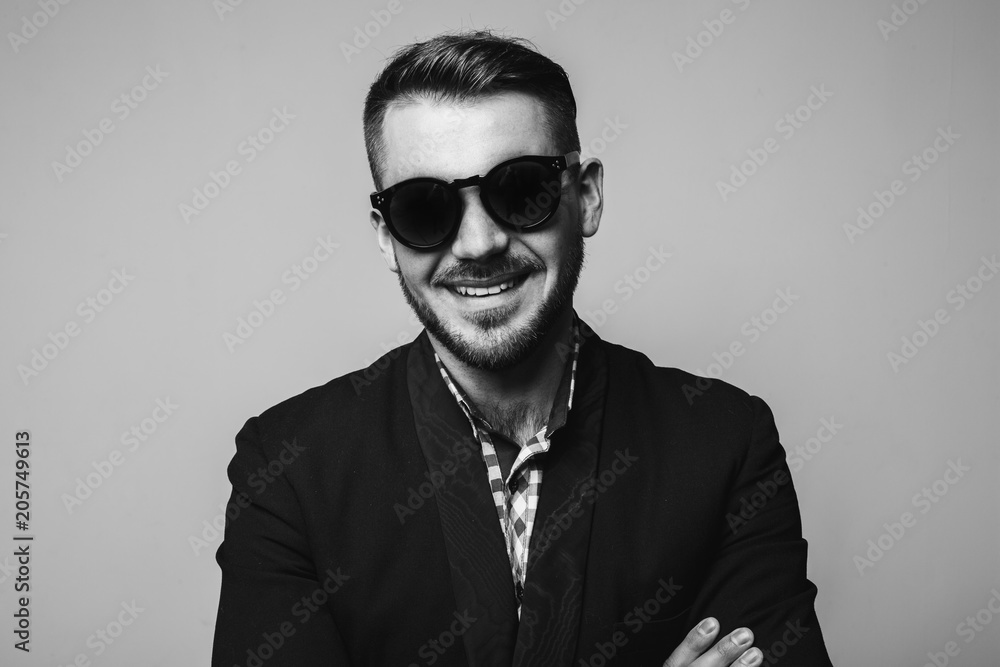 Close up portrait of attractive young business man in stylish black jacket, black sunglasses smiling isolated on grey background in the studio. Black and white shot Close up portrait of attractive young business man in stylish black jacket, black sunglasses smiling isolated on grey background in the studio. Black and white shot