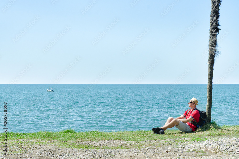 Man with backpack sits under a tall palm tree on background of sea and blue sky
