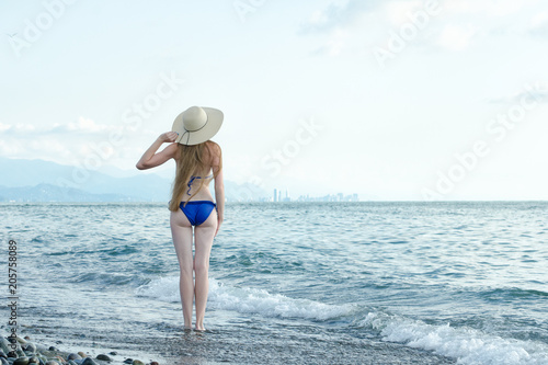 Girl in a swimsuit suit and hat is standing in sea waves. Back view