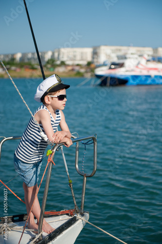 handsome little boy in sunglasses and captain cap on yacht board in sea port in summer vacation day