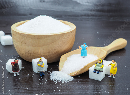 Miniature fat woman standing near wooden bowl and spoon with sugar cube on dark wood background, and thinking of weight loss and slim body. Healthy lifestyle concept.