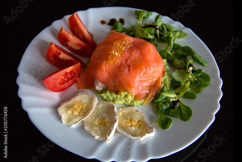 White plate with healthy meal on the black background, in the middle of plate is smoked salmon wrapped around avocado; topped with fish caviar, goat cheese; fresh tomato and lettuce on sides