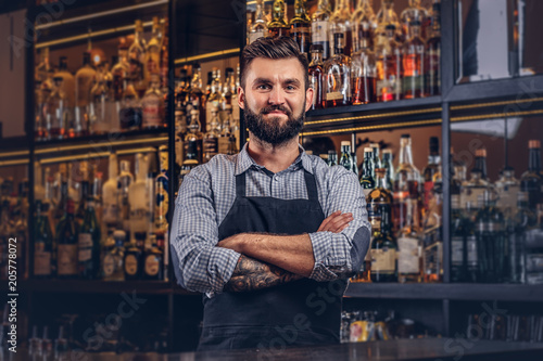 Stylish brutal bartender in a shirt and apron standing with crossed arms at bar counter background. photo