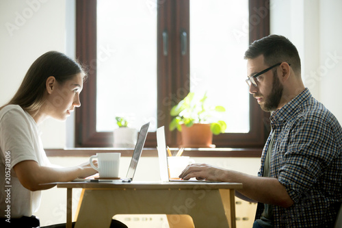 Obraz na plátně Work rivals feeling jealousy, envy, looking at each other behind laptops, competing in developing business strategy, staring defiantly with hate and distrust