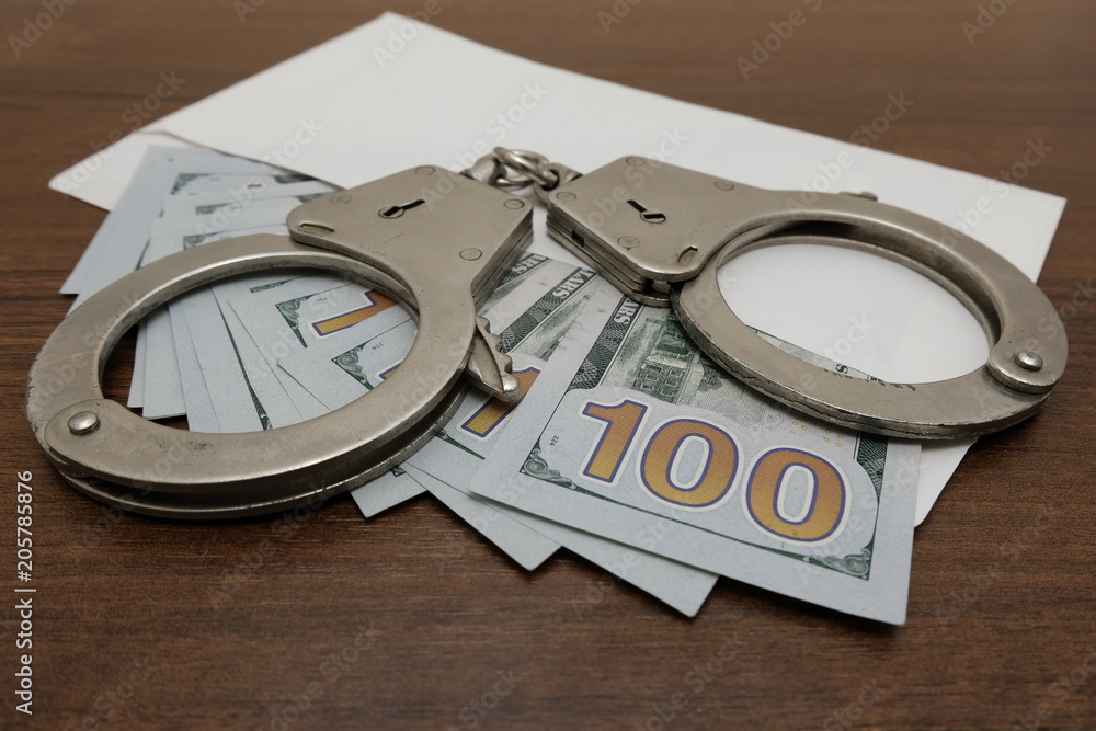 The handcuffs lie on an envelope in which are many hundred dollar bills. Brown wooden background. Concept violation of the law, corruption, financial fraud. The handcuffs lie on an envelope in which are many hundred dollar bills. Brown wooden background. Concept violation of the law, corruption, financial fraud.