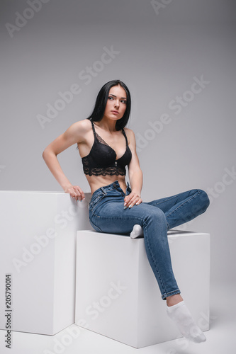 attractive sexy girl in black brassiere and jeans sitting on cube in photostudio