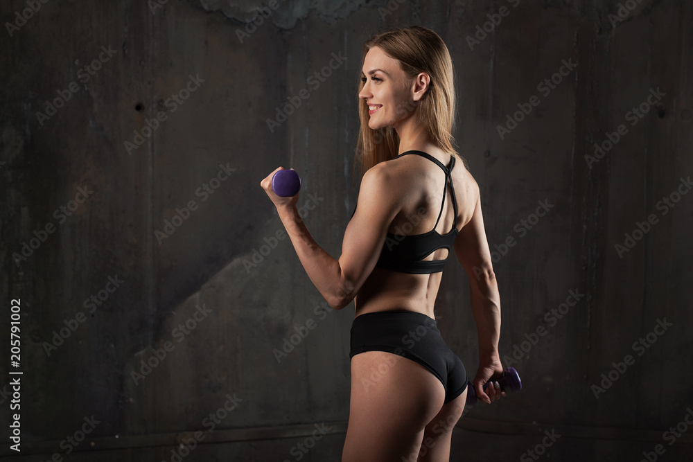 Image of muscular young female athlete wearing black sport wear stands with his back to the camera on dark background.
