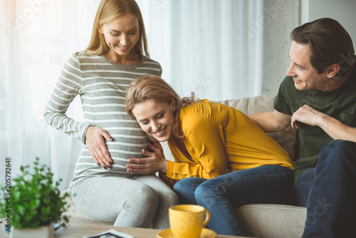 We are waiting for you. Excited young woman is leaning head on female abdomen and hearing to unborn child. Man is sitting near them and smiling. Surrogacy concept  photo