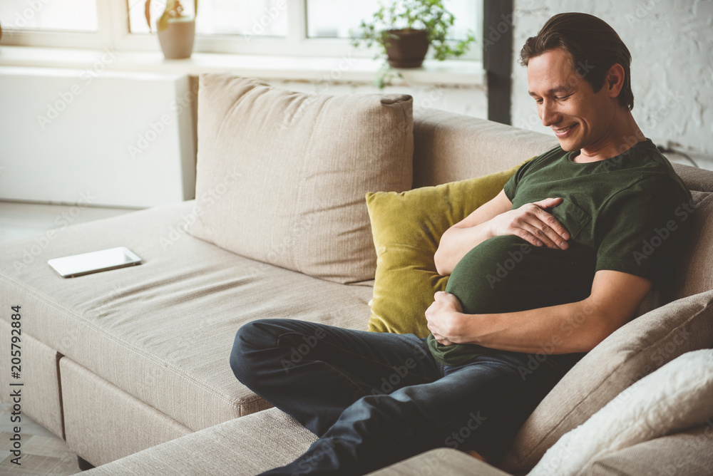Dreaming of future child. Cheerful young man is sitting on sofa as if he is pregnant. He is touching his belly with love and smiling. Copy space 