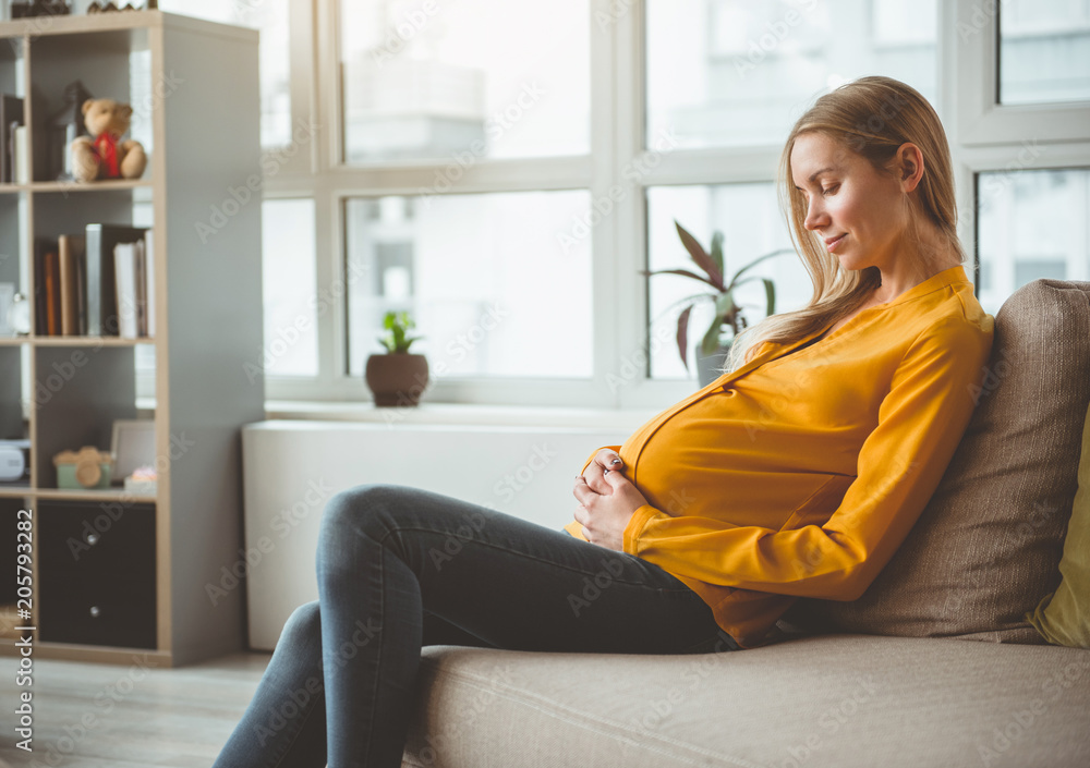 Calm young pregnant woman is sitting on couch with relaxation. She is embracing her belly with love 