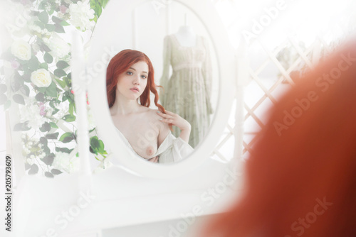 Tender portrait of a young dreamy redhead woman wearing beautiful vintage dress. She is sitting in front of the mirror with naked breast. photo