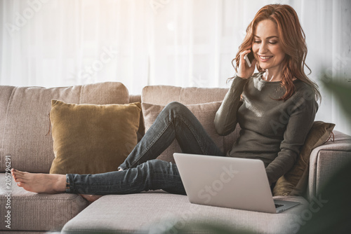 Full length portrait of carefree girl holding her mobile and communicating with smile and delight on couch at home. She is browsing information online and admiring free time in domestic atmosphere
