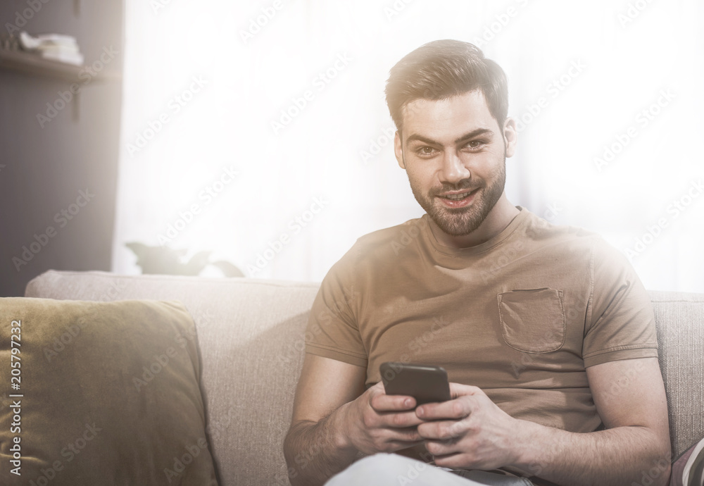 Good mood. Portrait of young attractive man with stubble is sitting on couch while looking at camera with smile. He is holding modern smartphone. Sunlight in background and copy space in the left side Good mood. Portrait of young attractive man with stubble is sitting on couch while looking at camera with smile. He is holding modern smartphone. Sunlight in background and copy space in the left side
