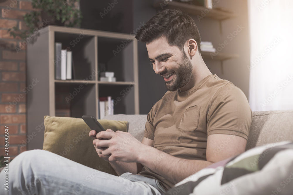Joyful news. Side view of cheerful young bristled man is sitting on comfortable couch at home and reading funny message on his modern mobile phone. He is looking at screen with smile Joyful news. Side view of cheerful young bristled man is sitting on comfortable couch at home and reading funny message on his modern mobile phone. He is looking at screen with smile