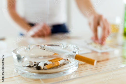 Cigarette in ashtray. On the background, woman playing with cards.