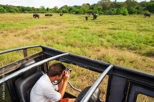 Photography Caucasian man in white shirt making photos of elephant herd during safari in nat