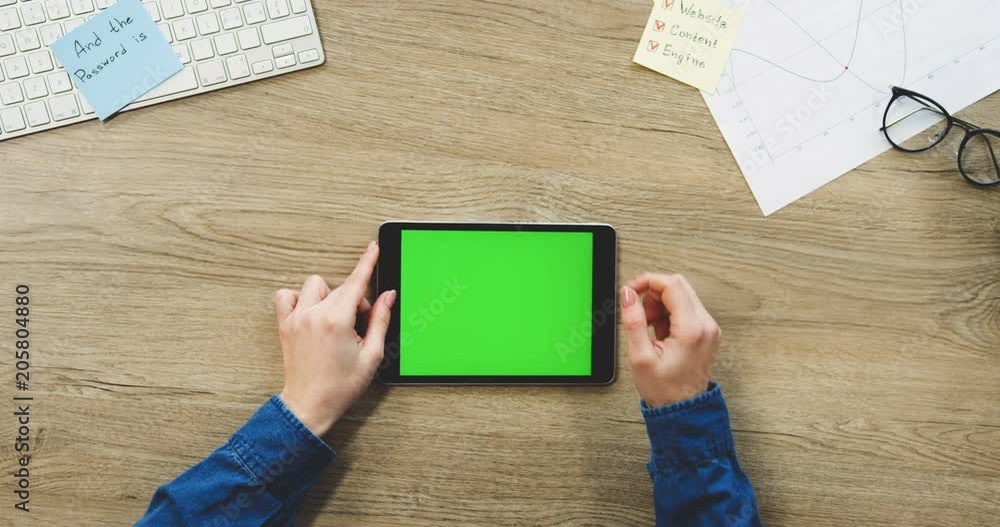 View from above on the woman's hands using the black tablet device with a green screen on the wooden office desk horizontally. Chroma key. Top view. Close up. View from above on the woman's hands using the black tablet device with a green screen on the wooden office desk horizontally. Chroma key. Top view. Close up.