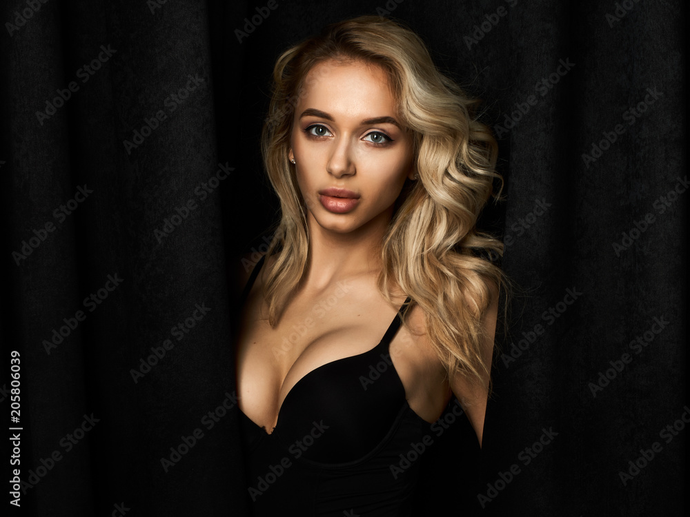 Fototapeta premium Beautiful blonde girl with long legs in black bodysuit against black velvet curtains in white sneakers.