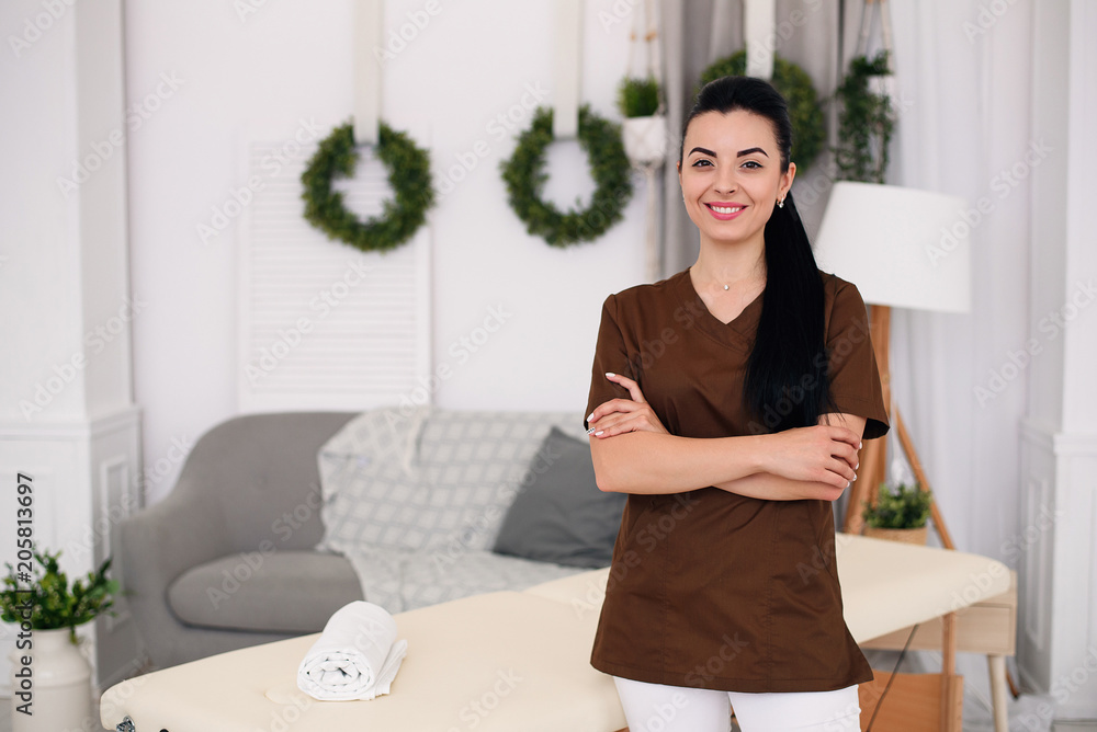 Smiling doctor looking at camera and standing behind massage table in modern medical office. Health care and medical concept. Smiling doctor looking at camera and standing behind massage table in modern medical office. Health care and medical concept.