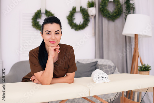 Slika na platnu Smiling female masseuse looking at camera and leaning on a massage table in a modern medical office