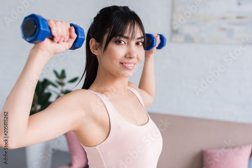 happy young woman working out with dumbbells at home and looking at camera