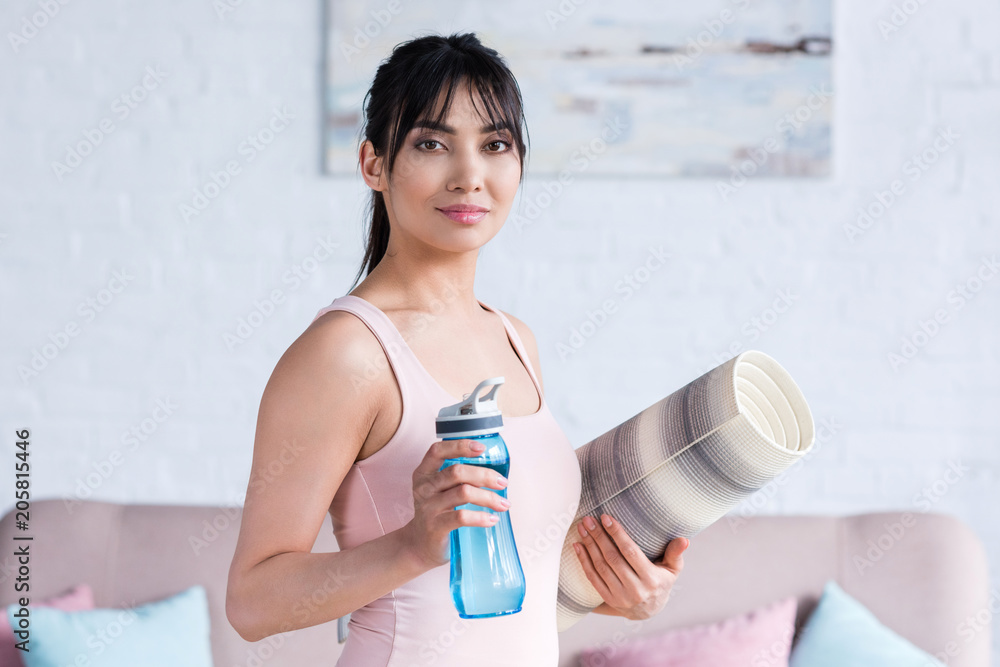 smiling young woman with rolled yoga mat and bottle of water looking at camera at home smiling young woman with rolled yoga mat and bottle of water looking at camera at home