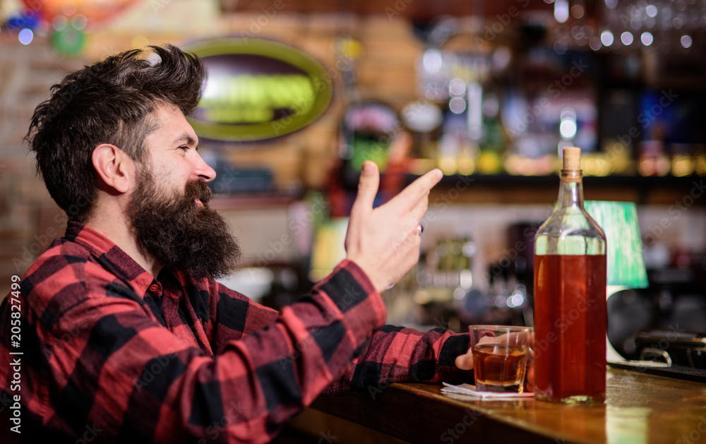 Hipster with beard ordered full bottle of alcohol. Relaxation concept. Man drinks whiskey or cognac. Man with happy face sits near bar counter. Guy spend leisure in bar, defocused background.