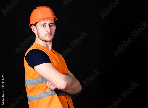 Strong builder concept. Man in helmet, hard hat hold arms crossed on chest, black background. Worker, contractor, builder on serious face with muscular biceps. Builder in helmet posing, copy space.
