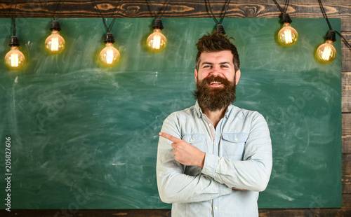Man with beard and mustache on happy smiling face stand in front of chalkboard. Bearded hipster in shirt, chalkboard on background copy space. Guy pointing with finger at board. Home task concept.