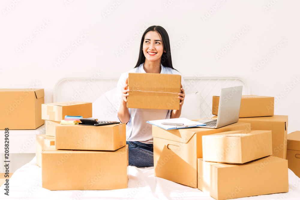 Young woman freelancer working and holding cardboard box on bed at home - SME business online and delivery concept Young woman freelancer working and holding cardboard box on bed at home - SME business online and delivery concept