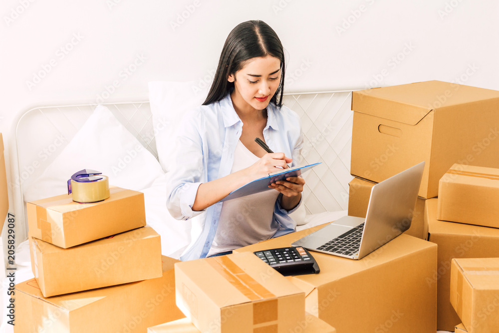 Young woman freelancer working and checklist and writing order with cardboard box on bed at home - SME business online and delivery concept Young woman freelancer working and checklist and writing order with cardboard box on bed at home - SME business online and delivery concept