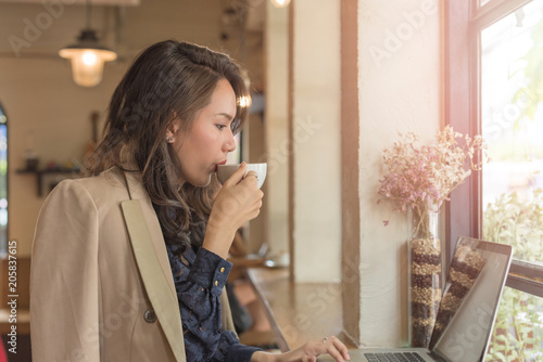 Young businesswoman sitting in coffee shop at table in front of laptop and drinking coffee. On background white brick wall and window. Girl shopping online, blogging, checking email.Online education.