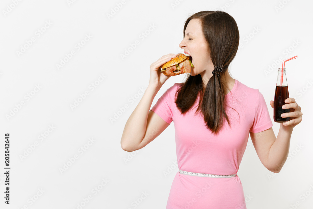 Fun young woman with tails, measuring tape at waist holding burger, cola in glass bottle isolated on white background. Proper nutrition or American classic fast food. Advertising area with copy space. Fun young woman with tails, measuring tape at waist holding burger, cola in glass bottle isolated on white background. Proper nutrition or American classic fast food. Advertising area with copy space.
