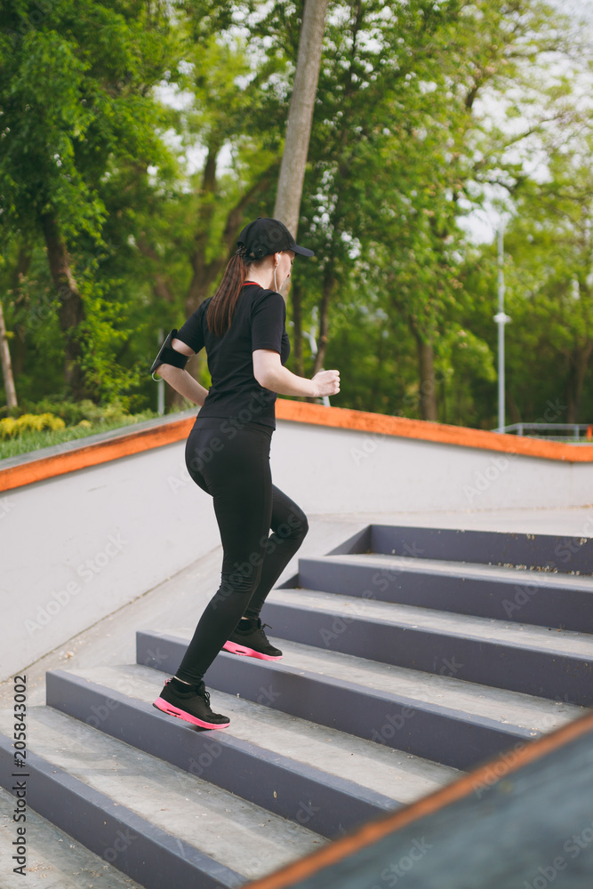 Young athletic beautiful brunette girl in black uniform, cap with headphones doing sport exercises, training and running, climbing on stairs in city park outdoors. Fitness, healthy lifestyle concept.