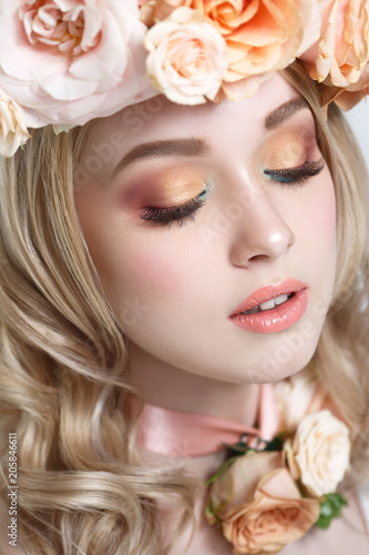 Fashion beauty portrait of a cute blonde girl in a wreath of tea roses and makeup in tone.