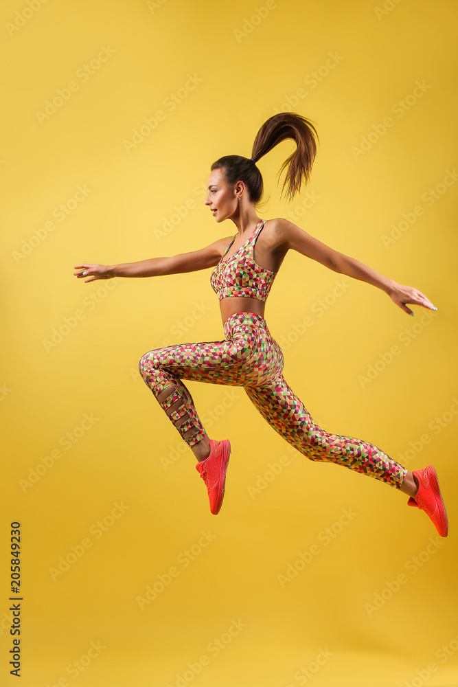Skinny girl with beautiful body, long legs, wearing colorful sportswear and sport shoes jumping high isolated on bright yellow background