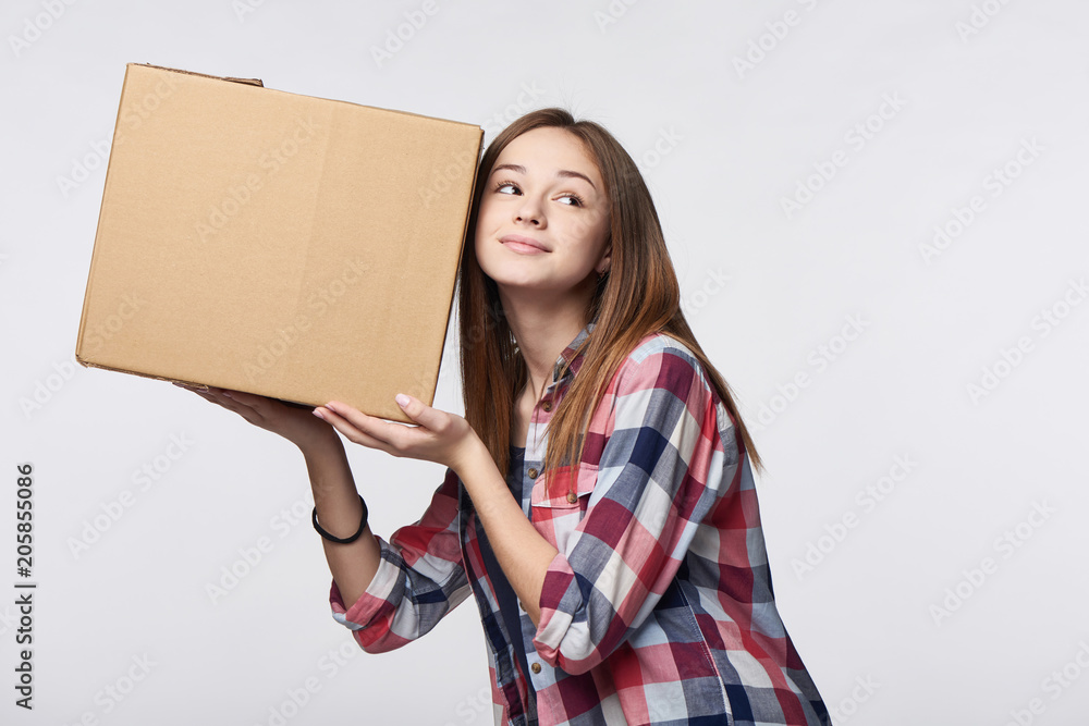 Delivery, relocation and unpacking. Smiling young woman holding cardboard box near her ear listening guessing what is inside, isolated Delivery, relocation and unpacking. Smiling young woman holding cardboard box near her ear listening guessing what is inside, isolated
