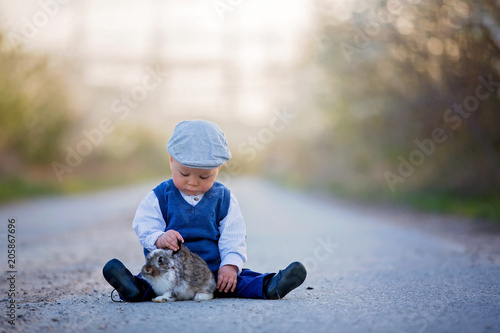 Adorable toddler boy, child playing with little bunny on a rural road with blooming trees photo