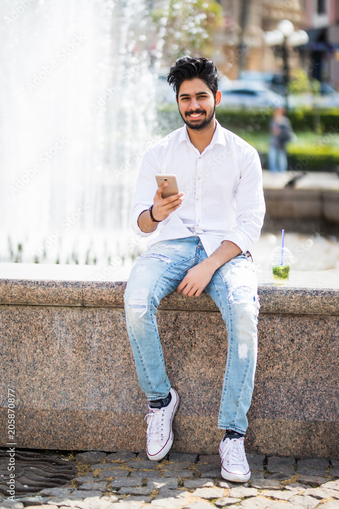 Happy handsome indian guy having walk in the city and typing sms sitting near fountain on the square Happy handsome indian guy having walk in the city and typing sms sitting near fountain on the square