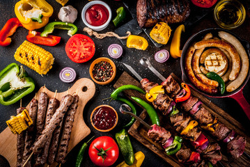 Assortment various barbecue food grill meat, bbq party fest - shish kebab, sausages, grilled meat fillet, fresh vegetables, sauces, spices, dark rusty concrete table, above copy space