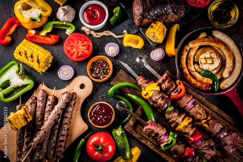Assortment various barbecue food grill meat, bbq party fest - shish kebab, sausages, grilled meat fillet, fresh vegetables, sauces, spices, dark rusty concrete table, above copy space photo